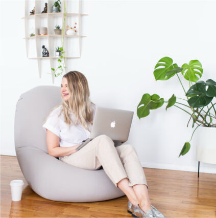 A woman sits on a bean bag chair, working on her laptop with a white wall, plants, and a shelf filled with small items in the background. Beside her, a coffee cup rests on the floor. Just behind her, a walkingpad is neatly folded against the wall.