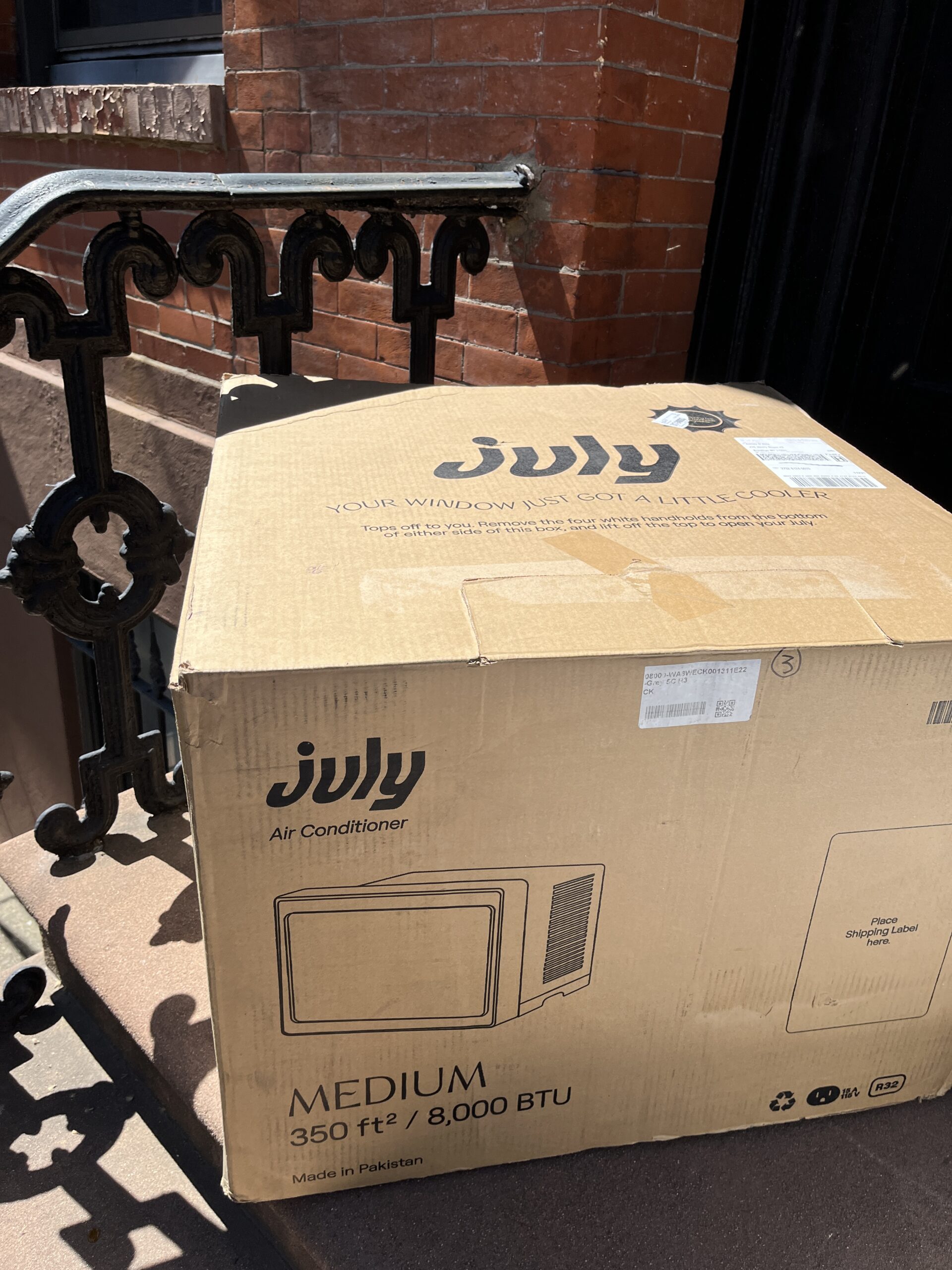 A large cardboard box labeled "July Air Conditioner" sits on a doorstep next to a wrought iron railing. The box is for a medium 8,000 BTU unit intended for 350 sq ft.