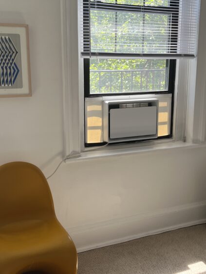 A yellow chair is next to a window with an air conditioning unit installed. The window has partially open blinds and there is a framed geometric artwork on the wall.