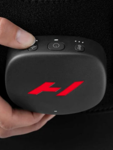 Close-up of a hand holding and adjusting a black device with a red "H" logo, featuring three buttons on top, reminiscent of high-end massage guns, attached to a black fabric strap.