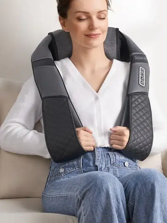 Woman sitting on a couch with a neck and shoulder massager draped over her shoulders, eyes closed, appearing relaxed. A tip displayed reads: "For maximum comfort, do not lean full body weight on the massager during use. Consider using massage guns for additional relief.