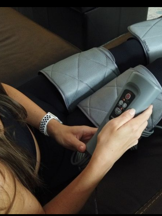 A woman is sitting on a couch, wearing leg compression sleeves and holding a control device, adjusting settings. 