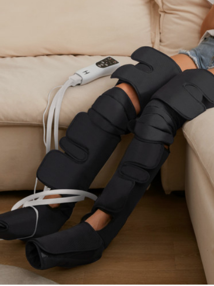 Person sitting on a beige couch with both legs wrapped in black compression therapy sleeves connected to a control unit for added muscle relief.