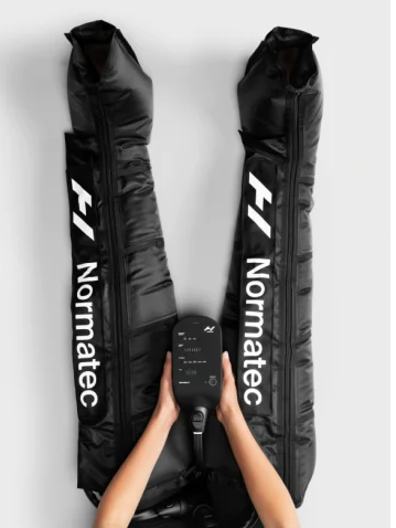 Person using a pair of Normatec compression leg sleeves with a control unit, complemented by massage guns for an enhanced recovery experience.