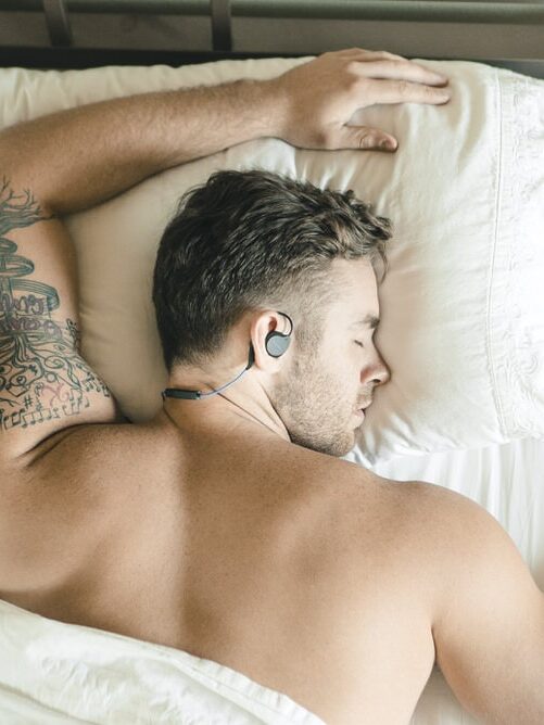 A tattooed man with earphones is sleeping on his side in a bed with white sheets and pillows, a massage gun resting by his nightstand.