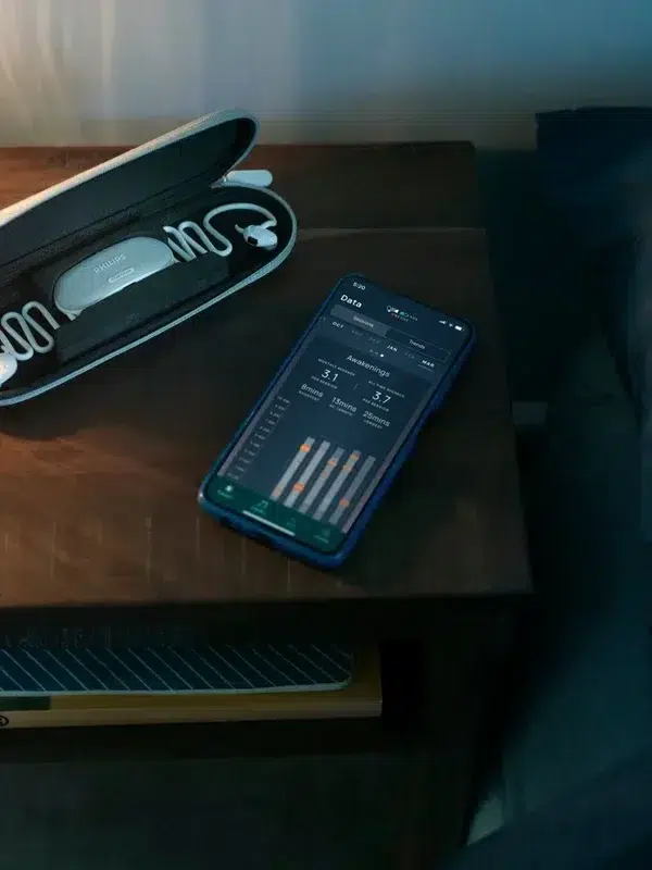 A smartphone displays an app next to a case containing earplugs, sleep aids, and a compact massage gun on a bedside table with a lamp.