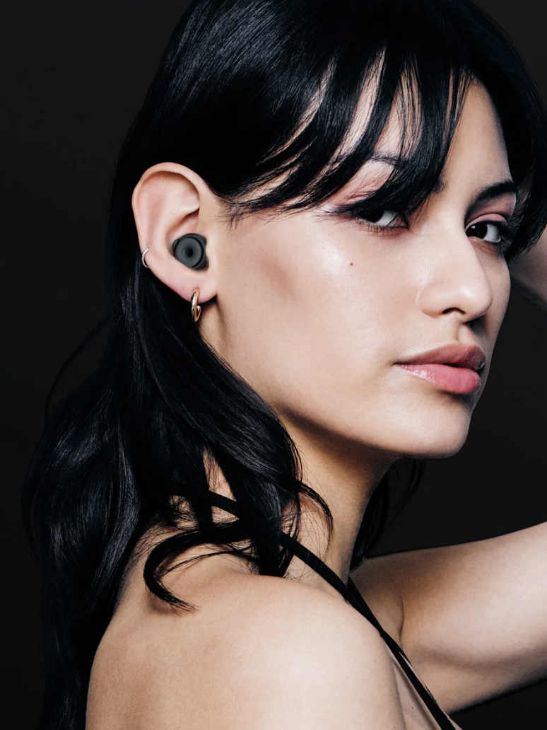 Person with long black hair and hoop earrings wearing wireless earbuds, holding a massage gun in one hand raised to their head, posing against a dark background.