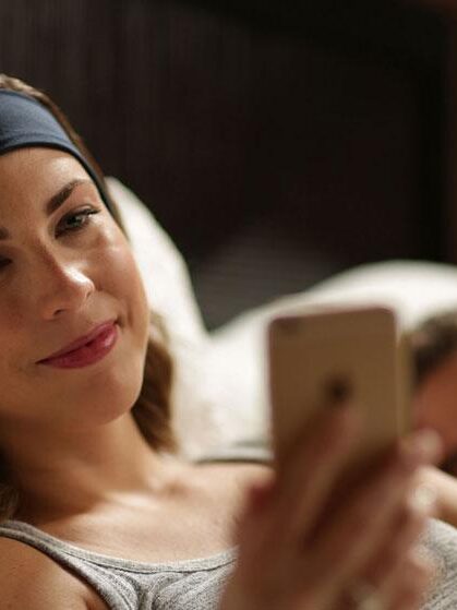 A woman wearing a headband lies on a bed using a smartphone, with a massage gun resting nearby, while a man sleeps beside her.