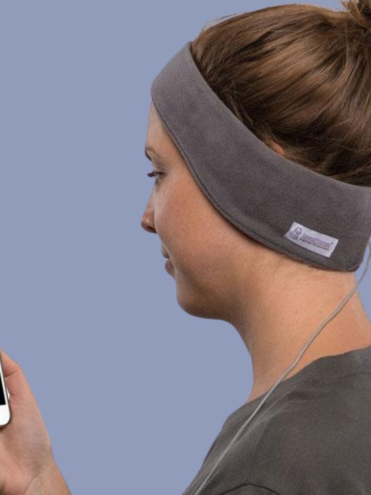 Person wearing a grey headband with built-in headphones, holding a smartphone and standing next to a new collection of sleek massage guns, all set against a plain light blue background.