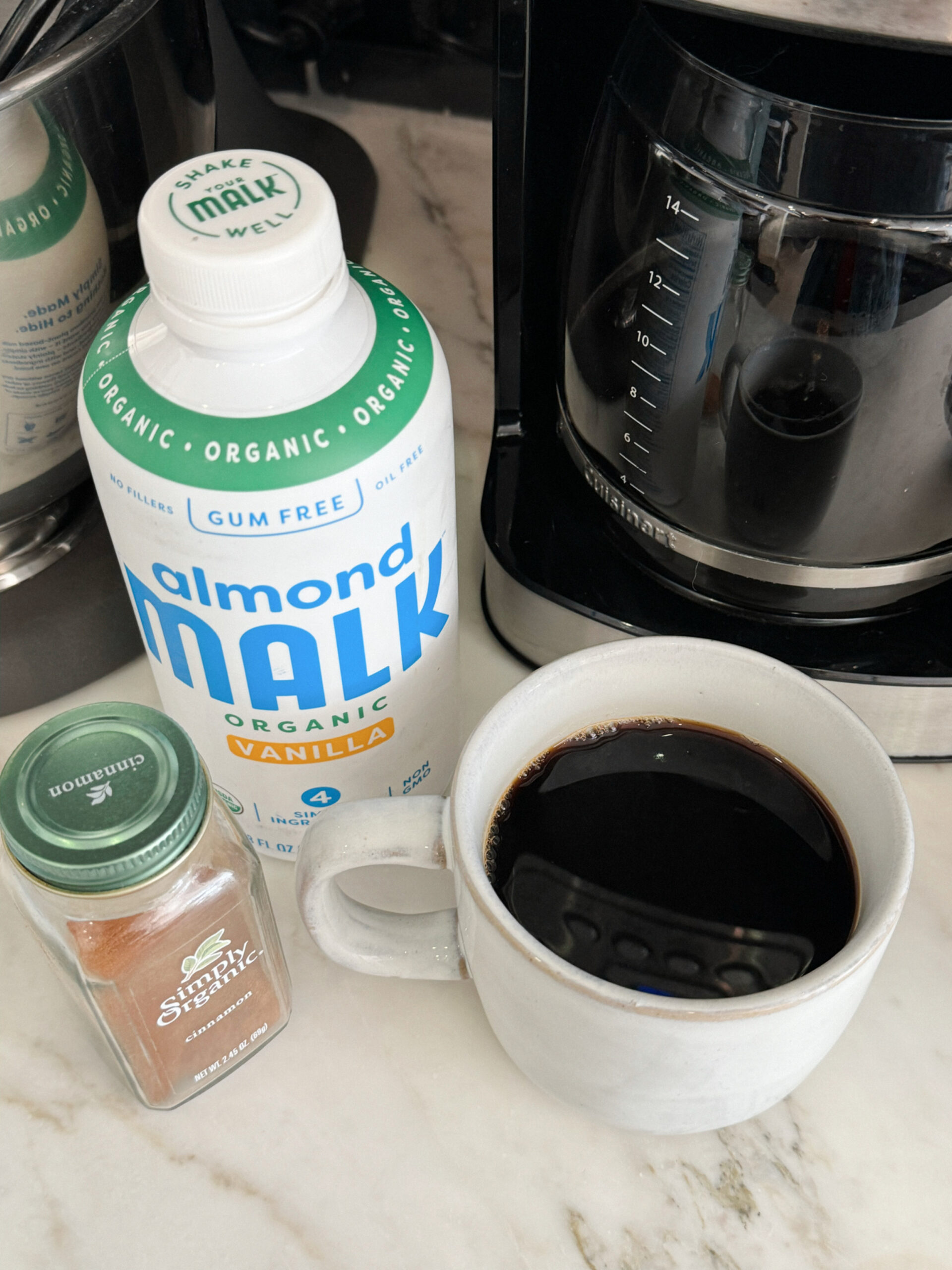 Cup of black coffee, container of vanilla almond milk, and a small jar of cinnamon placed next to a coffee maker on a kitchen counter.