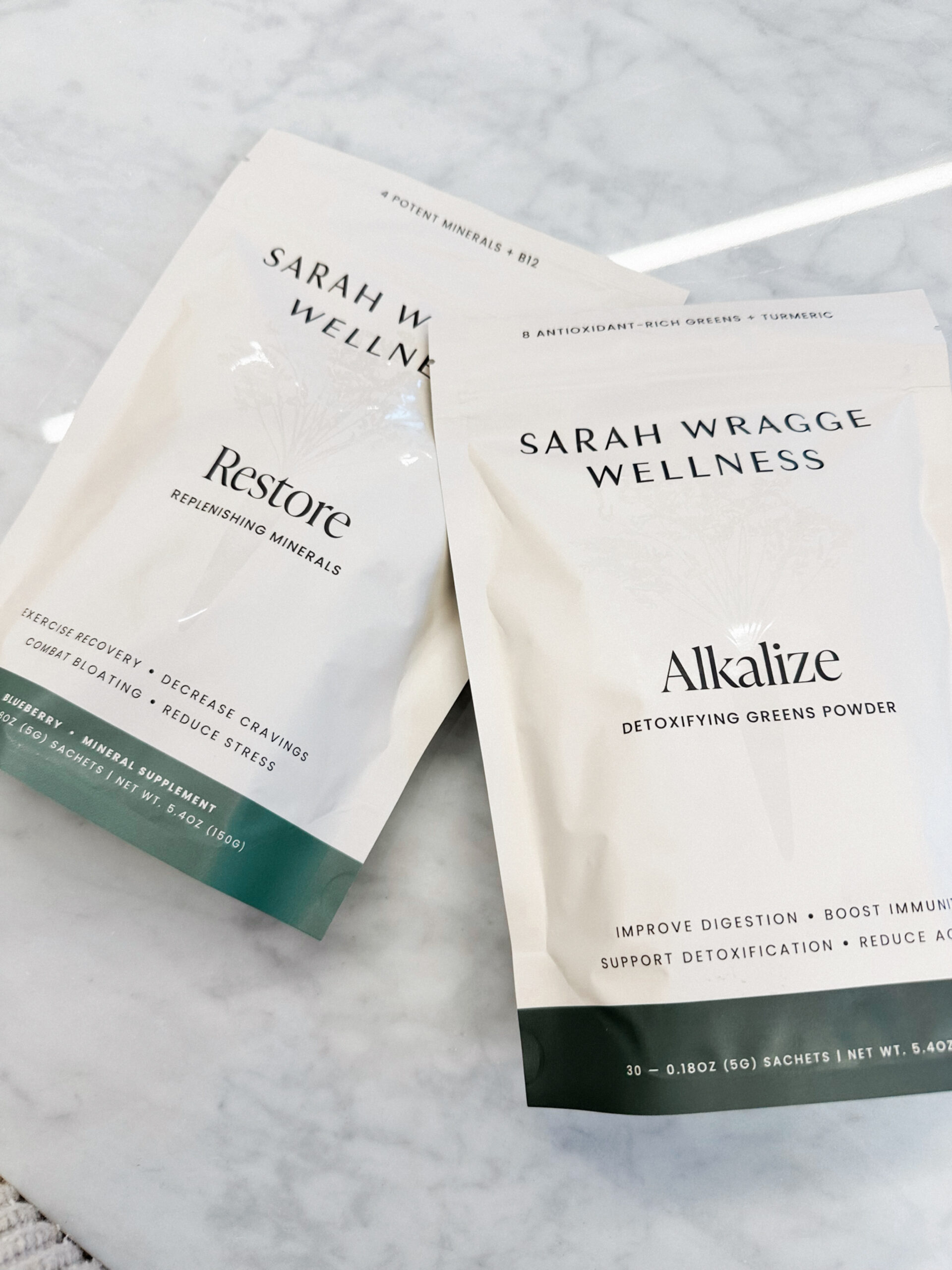 Two white packets of Sarah Wragge Wellness products on a marble surface. One packet is labeled "Restore" and the other "Alkalize".