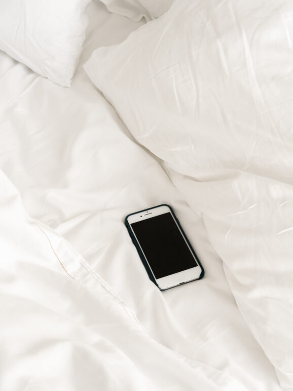 A smartphone with a black case is lying on a white bed with crumpled sheets and pillows.