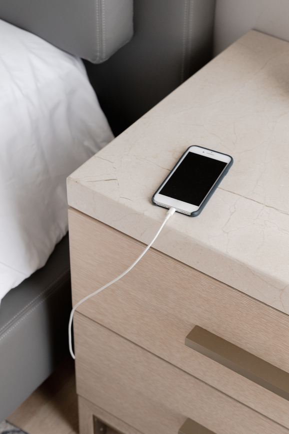 A smartphone connected to a charger rests on a light-colored nightstand next to a bed.