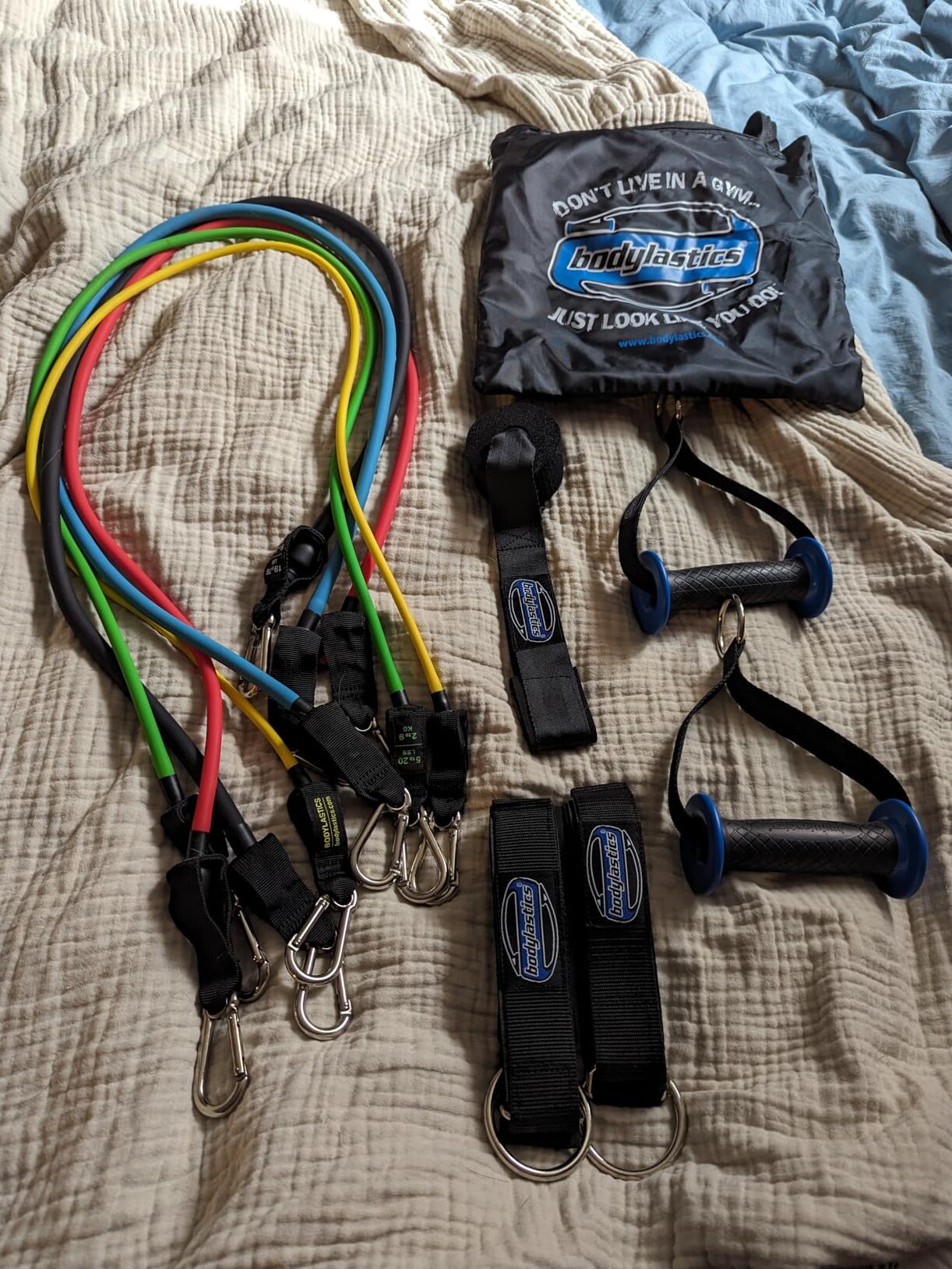 A set of colorful resistance bands, handles, and straps with clips laid on a bed next to a Bodylastics branded bag, accompanied by a sleek massage gun.