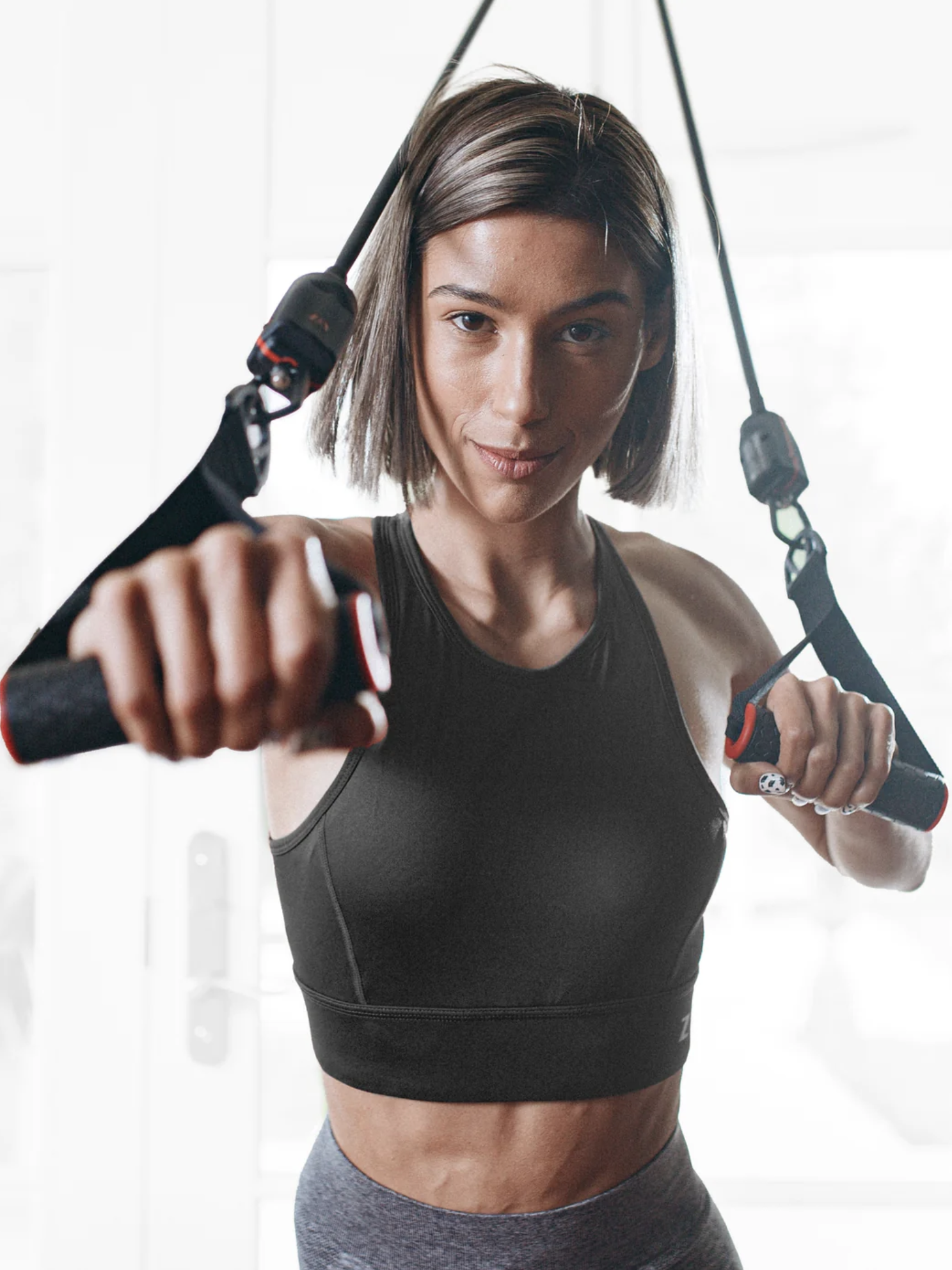 A woman in athletic wear uses suspension training straps while working out indoors, and a massage gun lies nearby for post-workout recovery.