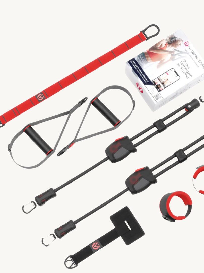 A suspension training set with red and black straps, handles, clips, a box of instructions, and a complementary massage gun is displayed on a white background.