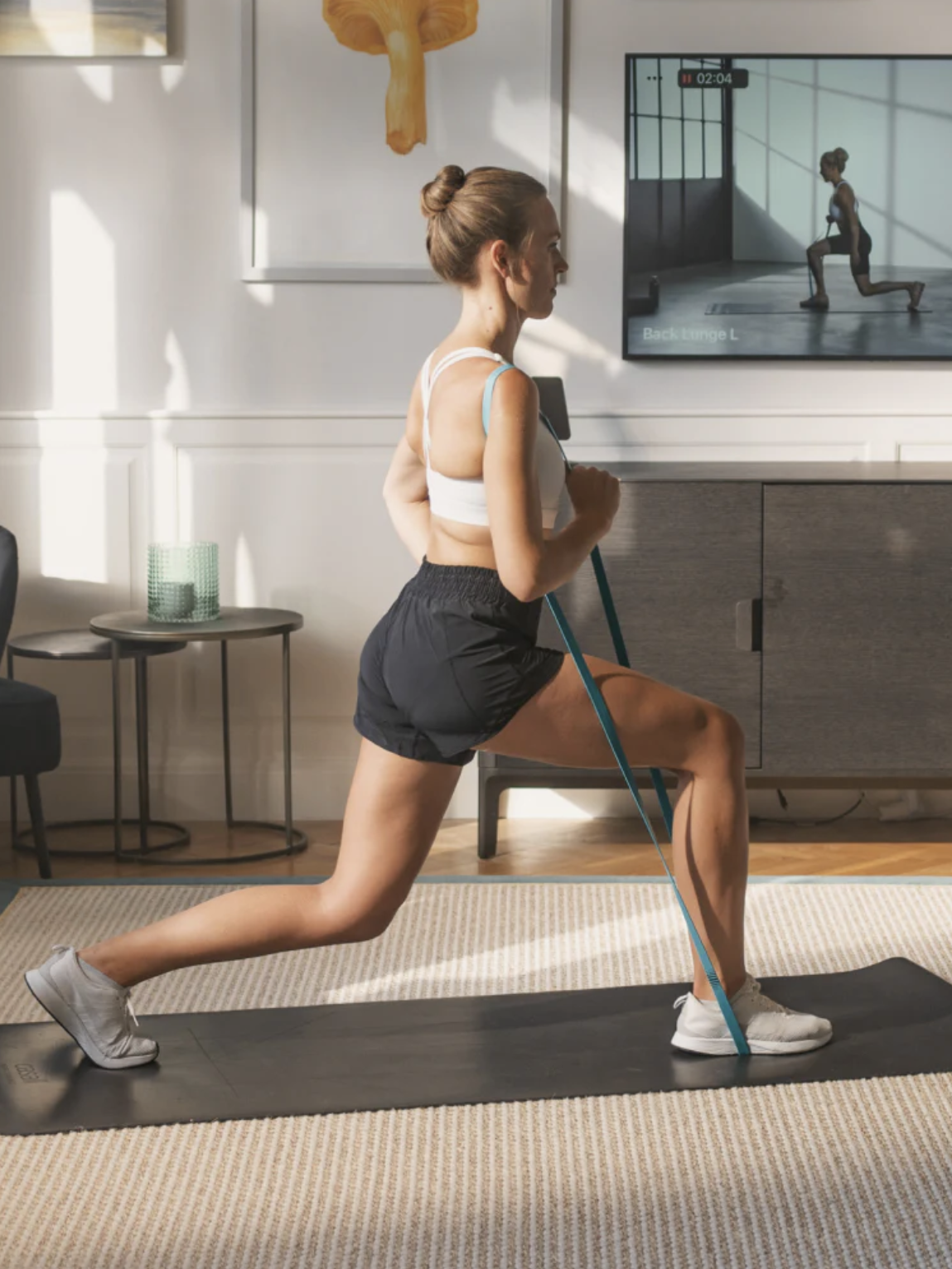 A woman is performing a resistance band exercise in a living room. She is in a lunge position while pulling the band with her arms. There is an exercise-related video playing on a TV in the background, and a massage gun rests nearby, ready to soothe her muscles post-workout.