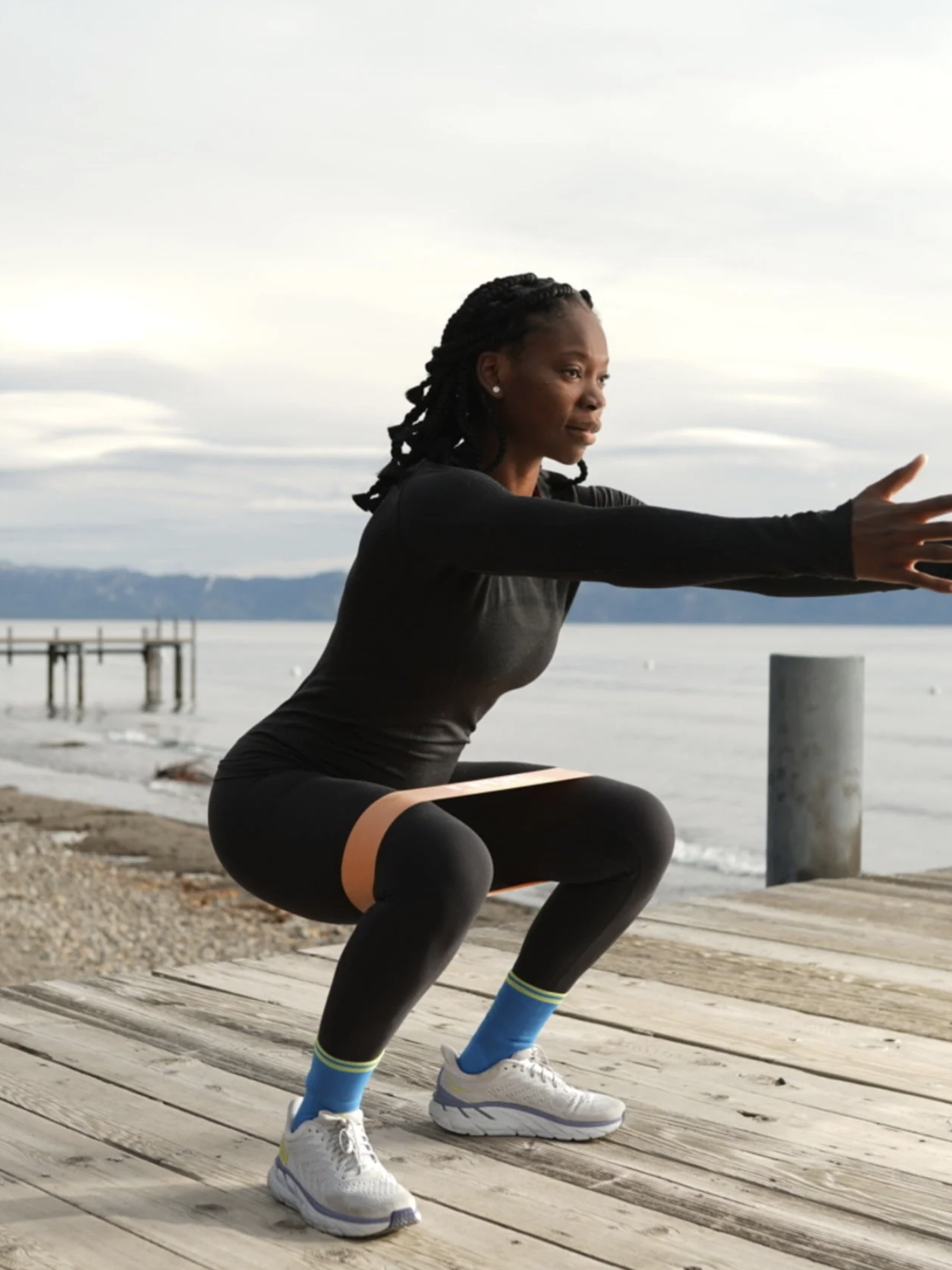 A person in athletic wear performs a squat exercise on a wooden boardwalk near a body of water with overcast skies, while a massage gun waits patiently nearby for post-workout recovery.