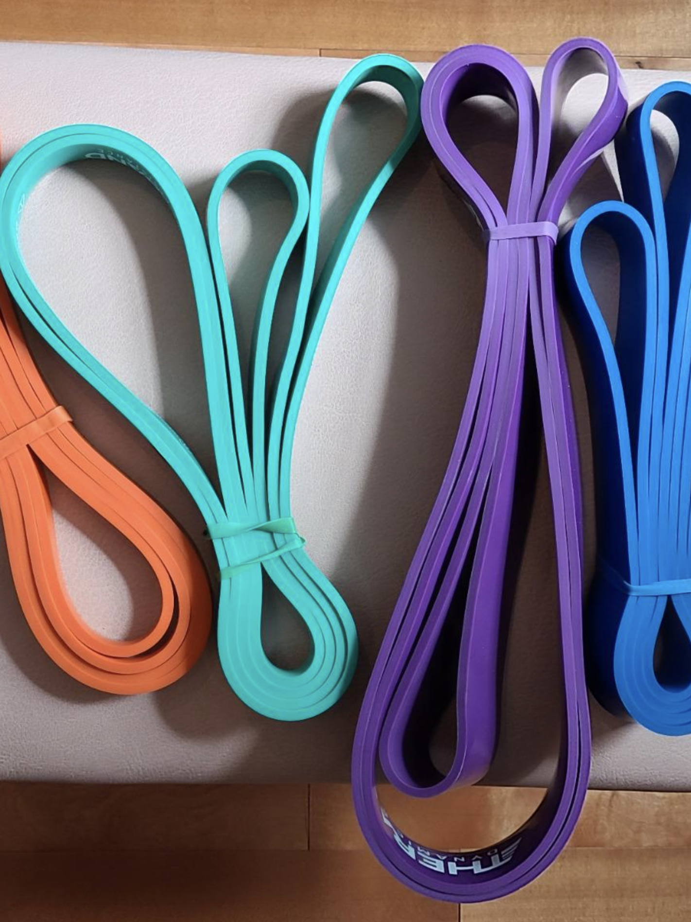 Four looped resistance bands in orange, green, purple, and blue are neatly placed on a light-colored surface alongside a couple of sleek massage guns.