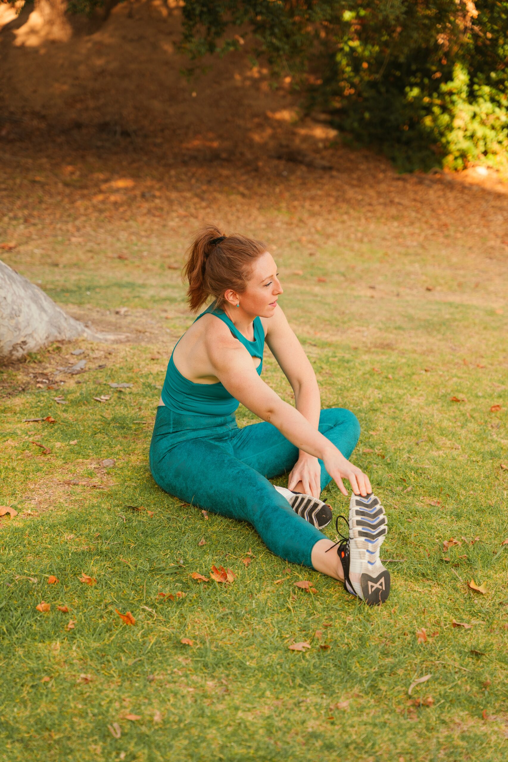 A woman in a teal workout outfit sits on the grass in a park, stretching her legs. She appears to be in the midst of a fitness routine.