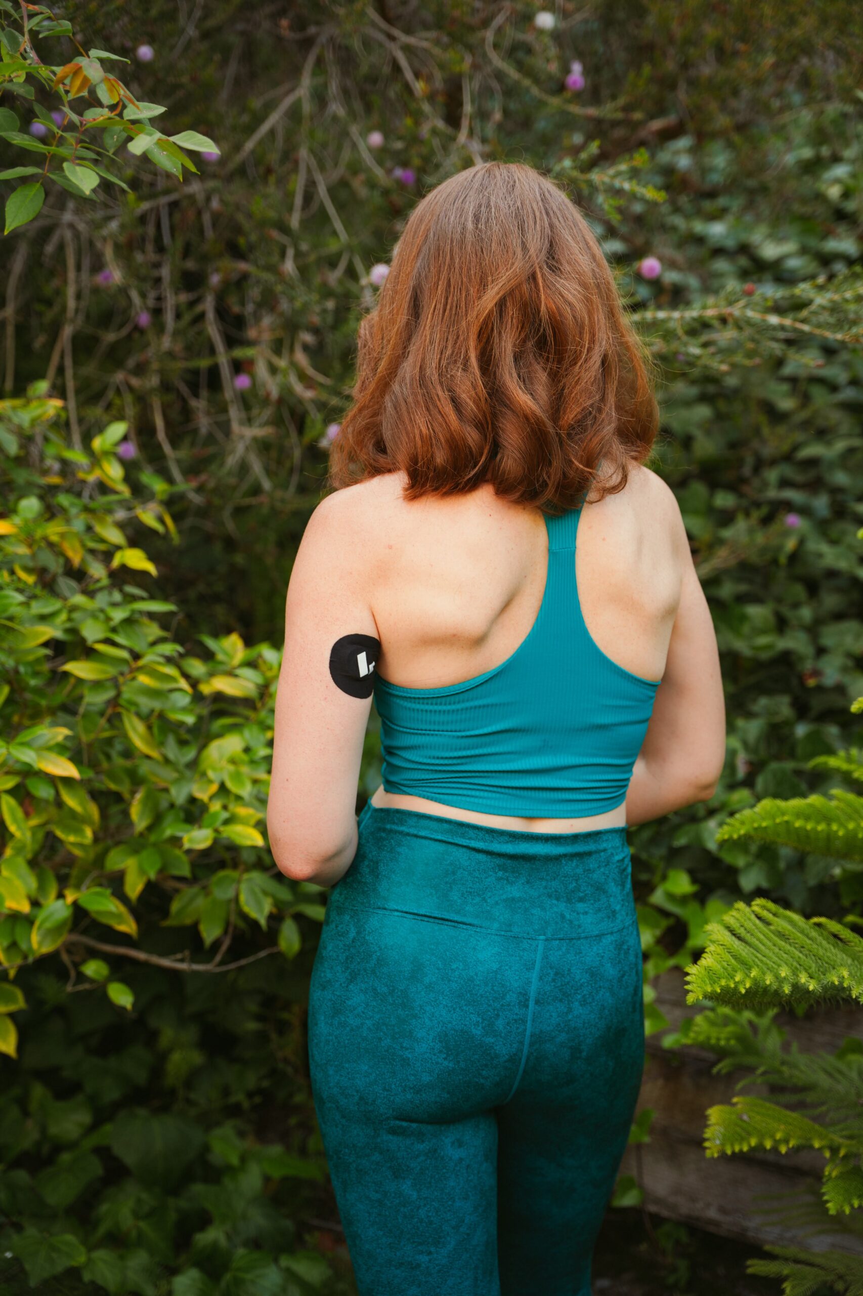 A person with medium-length auburn hair wearing a teal sports top and leggings stands outdoors with their back facing the camera, surrounded by lush green foliage.