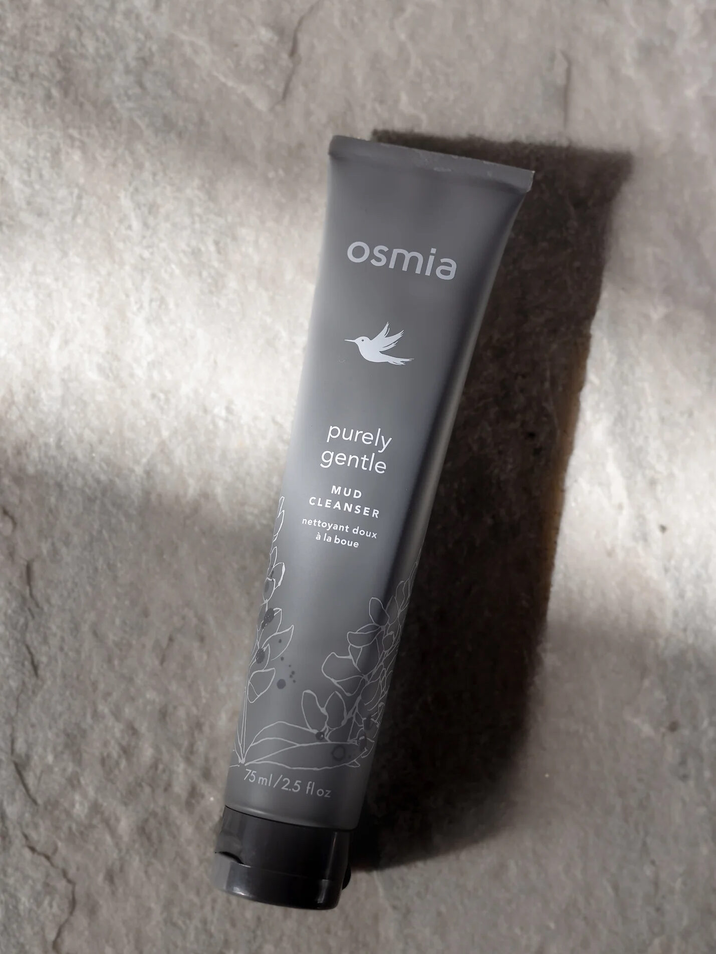 A gray tube of Osmia Purely Gentle Mud Cleanser with floral designs and product information on a stone surface, casting a shadow on the textured background.