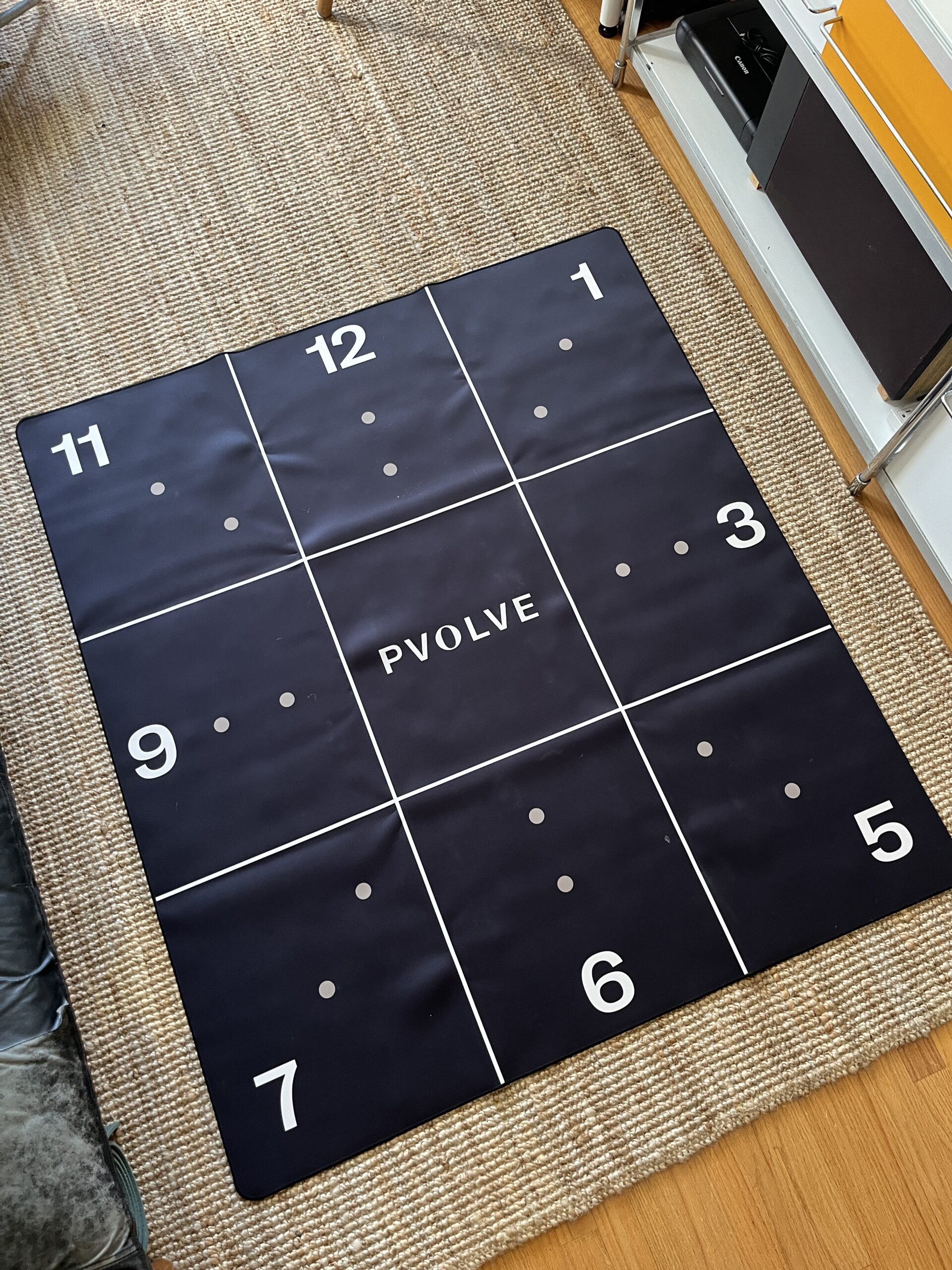 A black exercise mat with numbered sections from 1 to 12 and "P. VOLVE" printed in the center, placed on a woven rug in a room.