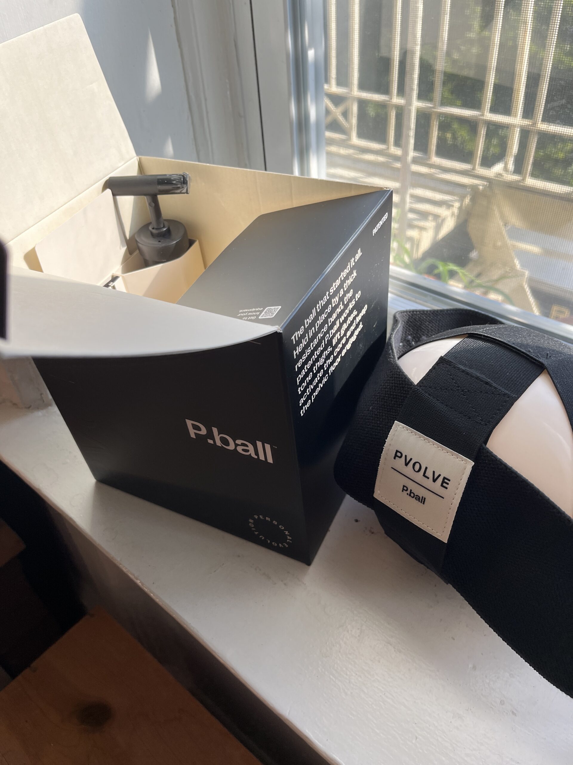 A P.volve P.ball exercise device partially removed from its black and white packaging is placed near a window with sunlight streaming in.