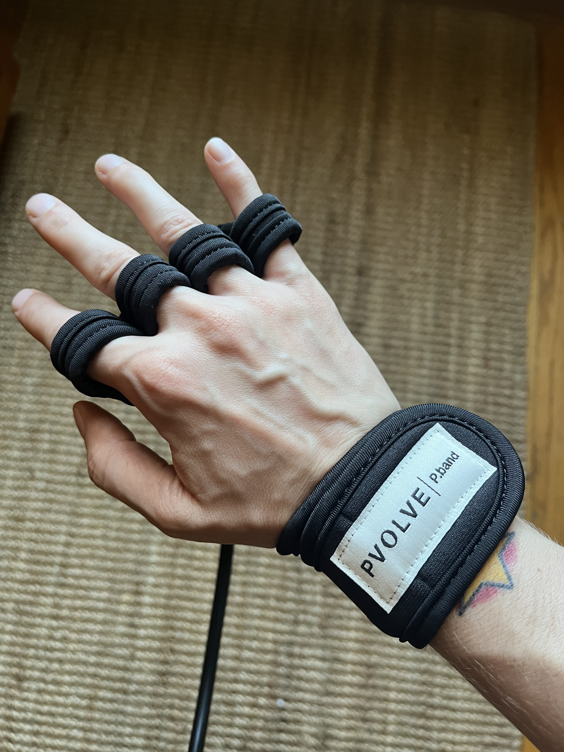 A hand wearing a black fitness resistance band device labeled "Pvolve" with multiple loops around the fingers and wrist, positioned over a textured surface.