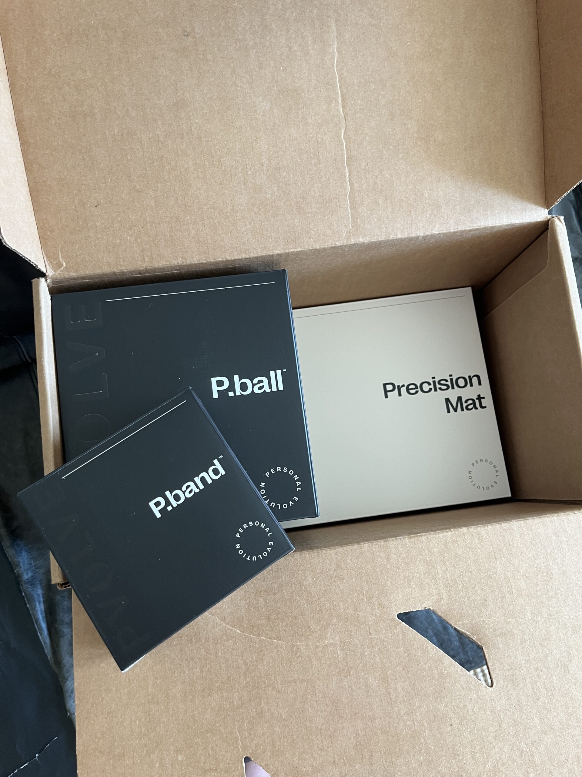 Opened cardboard box containing fitness equipment: a P.ball, a P.band, a Precision Mat, and an additional item.