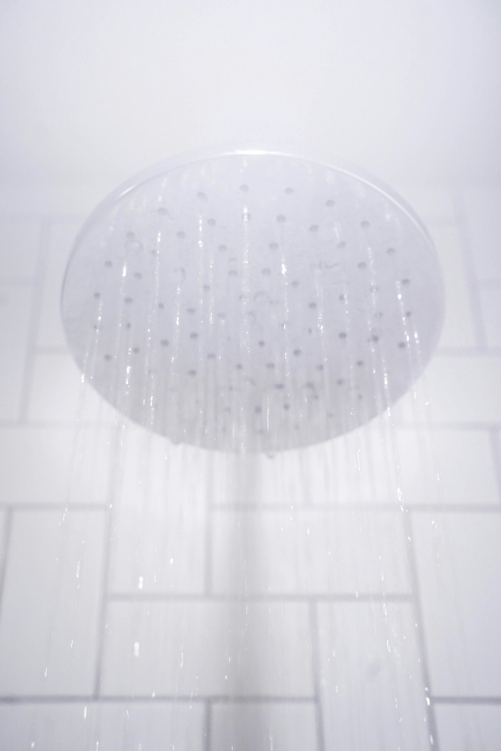 A round showerhead releasing water, set against a white tiled background.