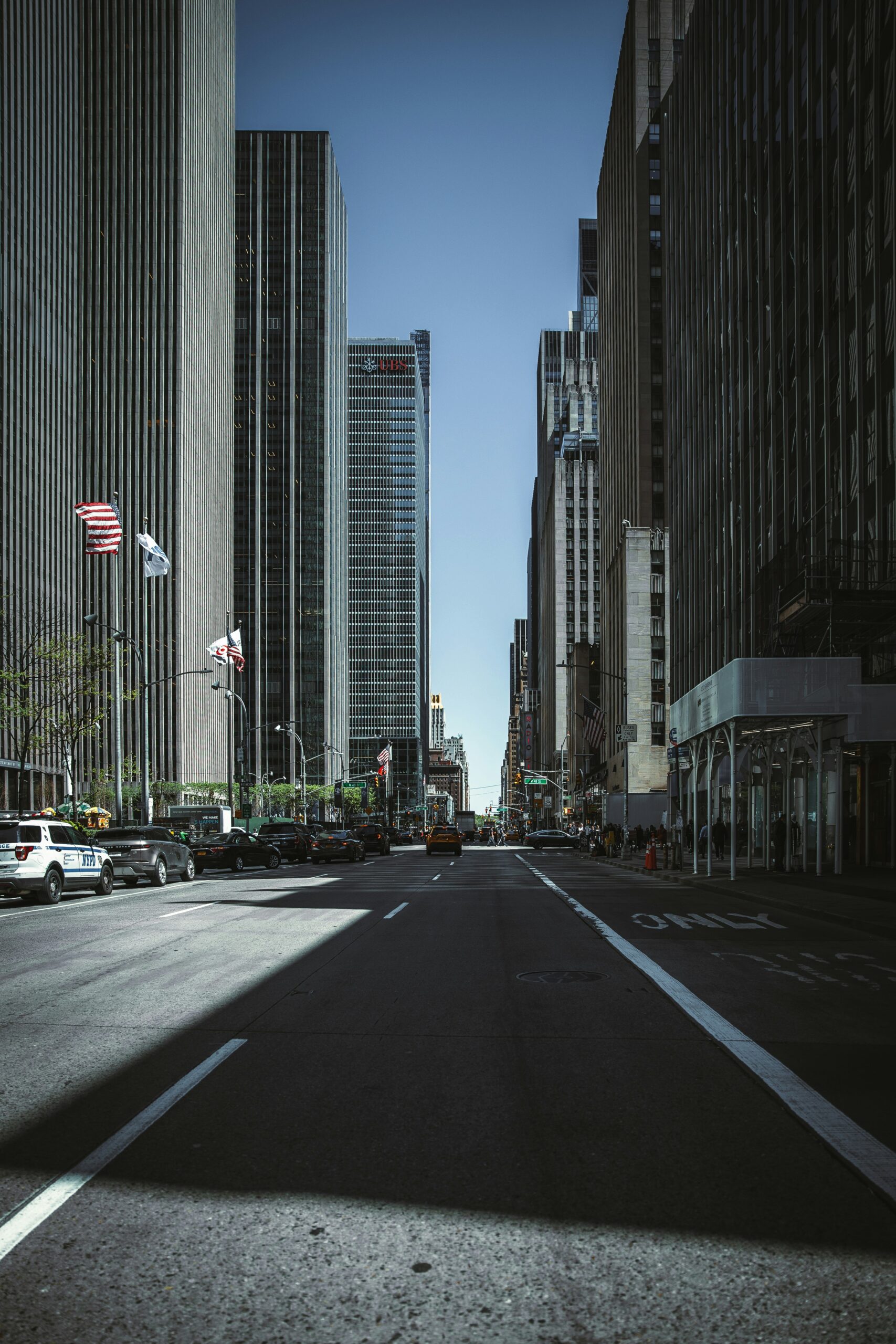 A wide city street lined with tall skyscrapers under a clear sky, with several flags on the left side and light traffic.