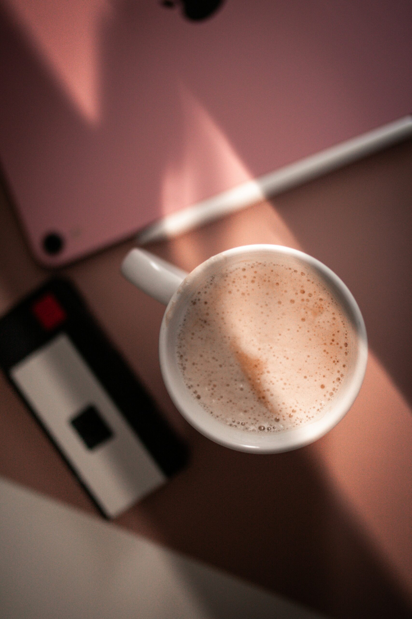 A cup of frothy coffee sits on a table beside a smartphone and an electronic device. Sunlight casts shadows across the scene.