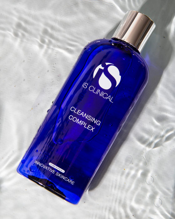 A blue bottle of IS Clinical Cleansing Complex with a silver cap is laying on a water surface. The label reads "Innovative Skincare.