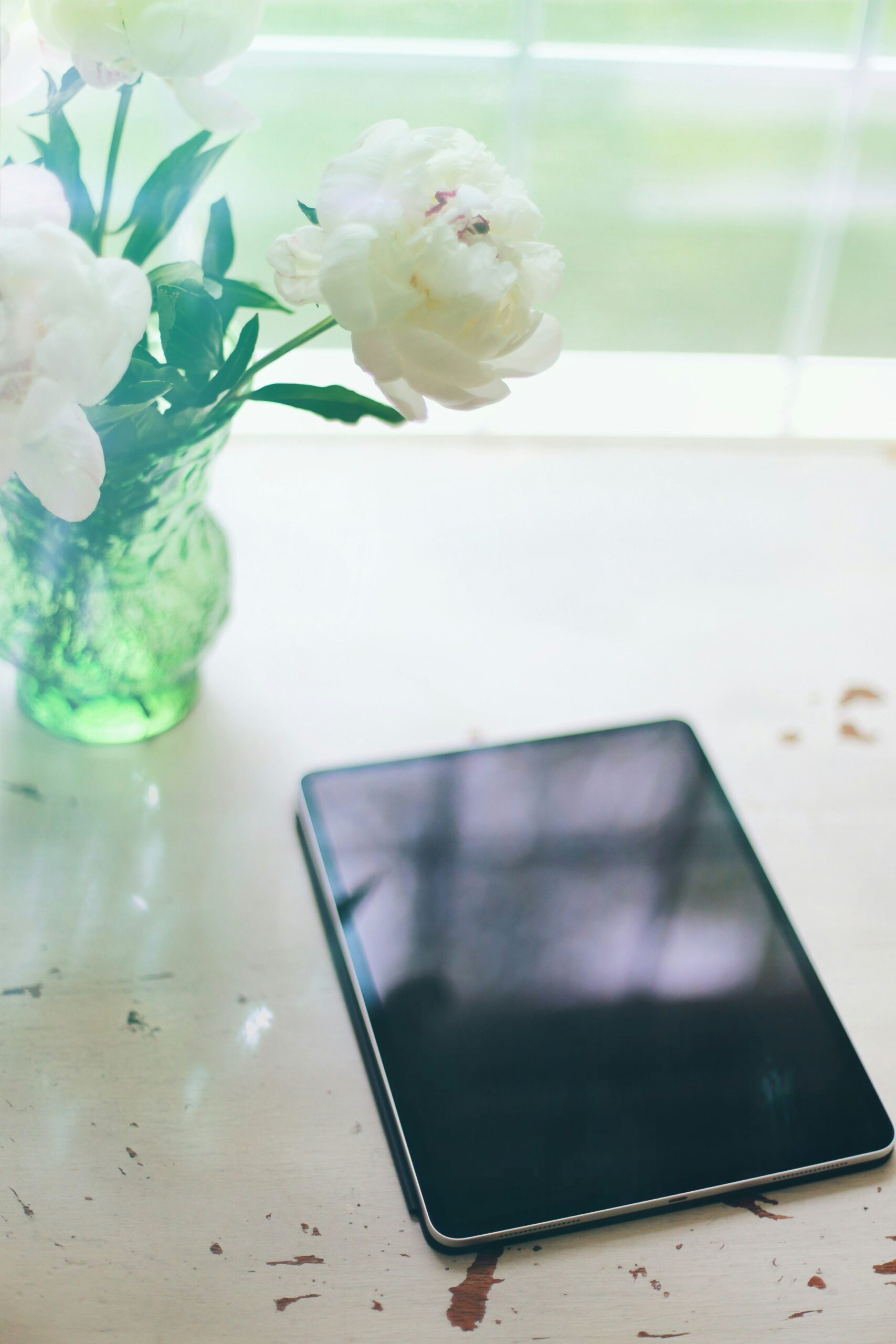 A tablet lies on a white table next to a green vase containing white flowers.