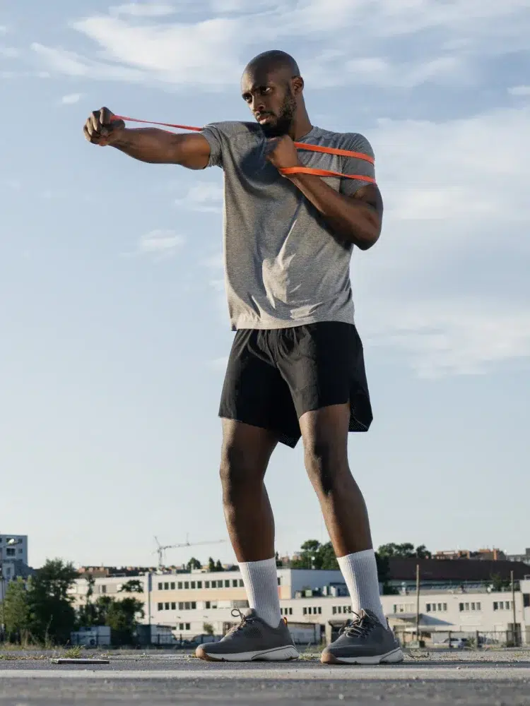 A man in athletic wear is exercising outdoors with a resistance band, incorporating massage guns into his routine. He stands on a rooftop with buildings and a clear sky in the background.