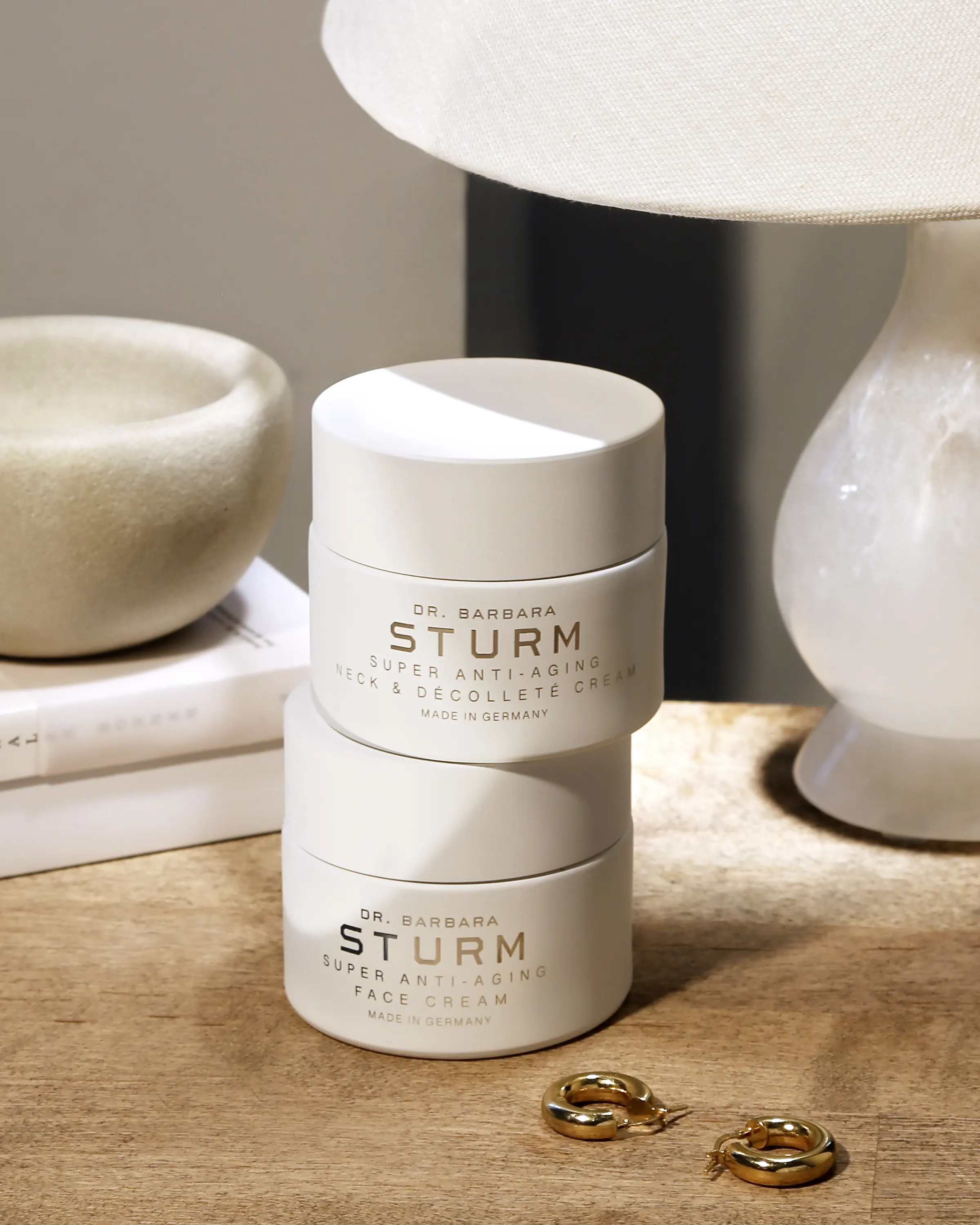 Two jars of Dr. Barbara Sturm skincare cream are stacked on a wooden surface next to a lamp, a pair of earrings, and a decorative bowl.