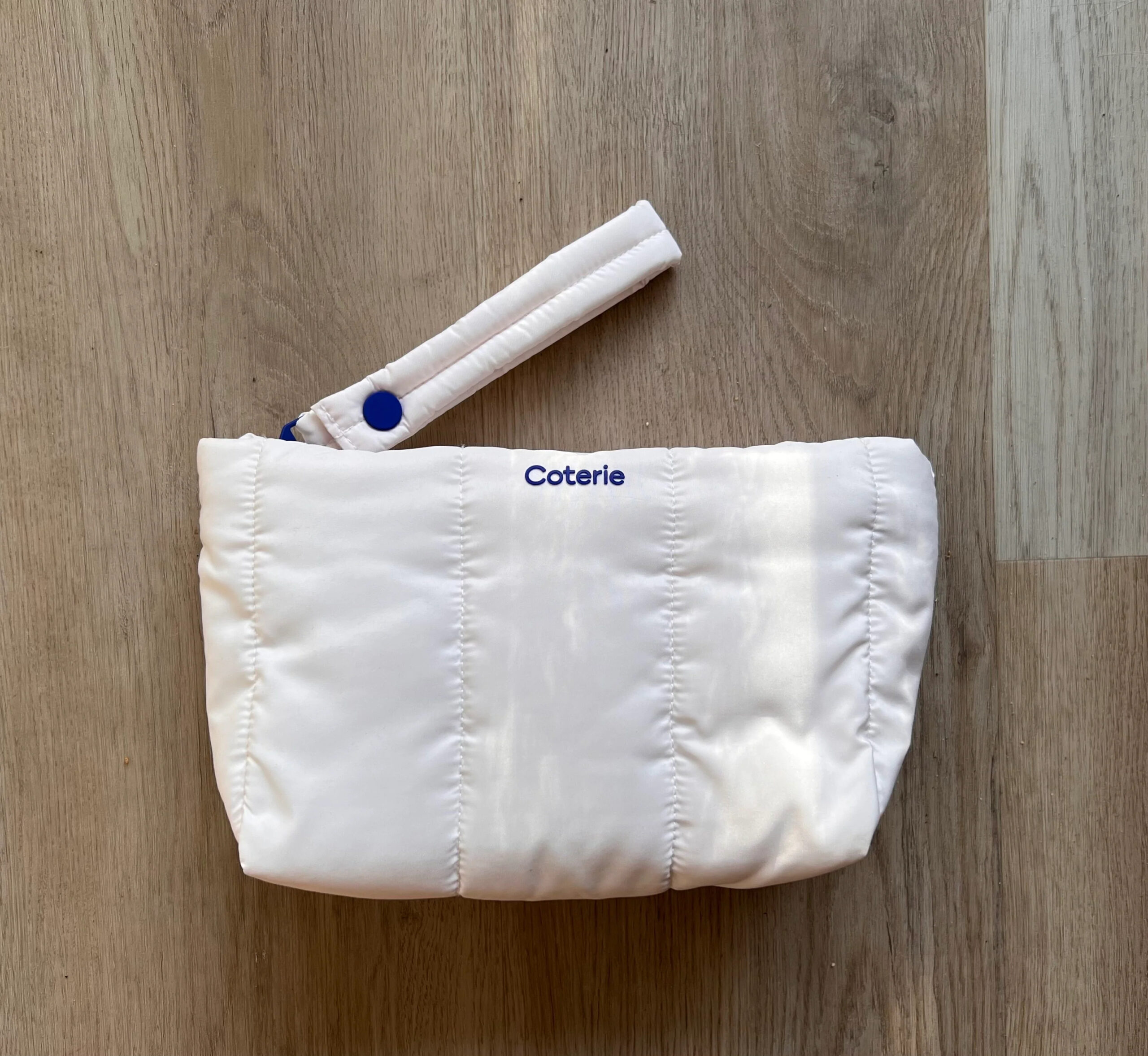 A small white quilted pouch with a handle and blue snap button, featuring the brand name "Coterie" embroidered in blue near the top. The pouch is placed on a wooden surface.