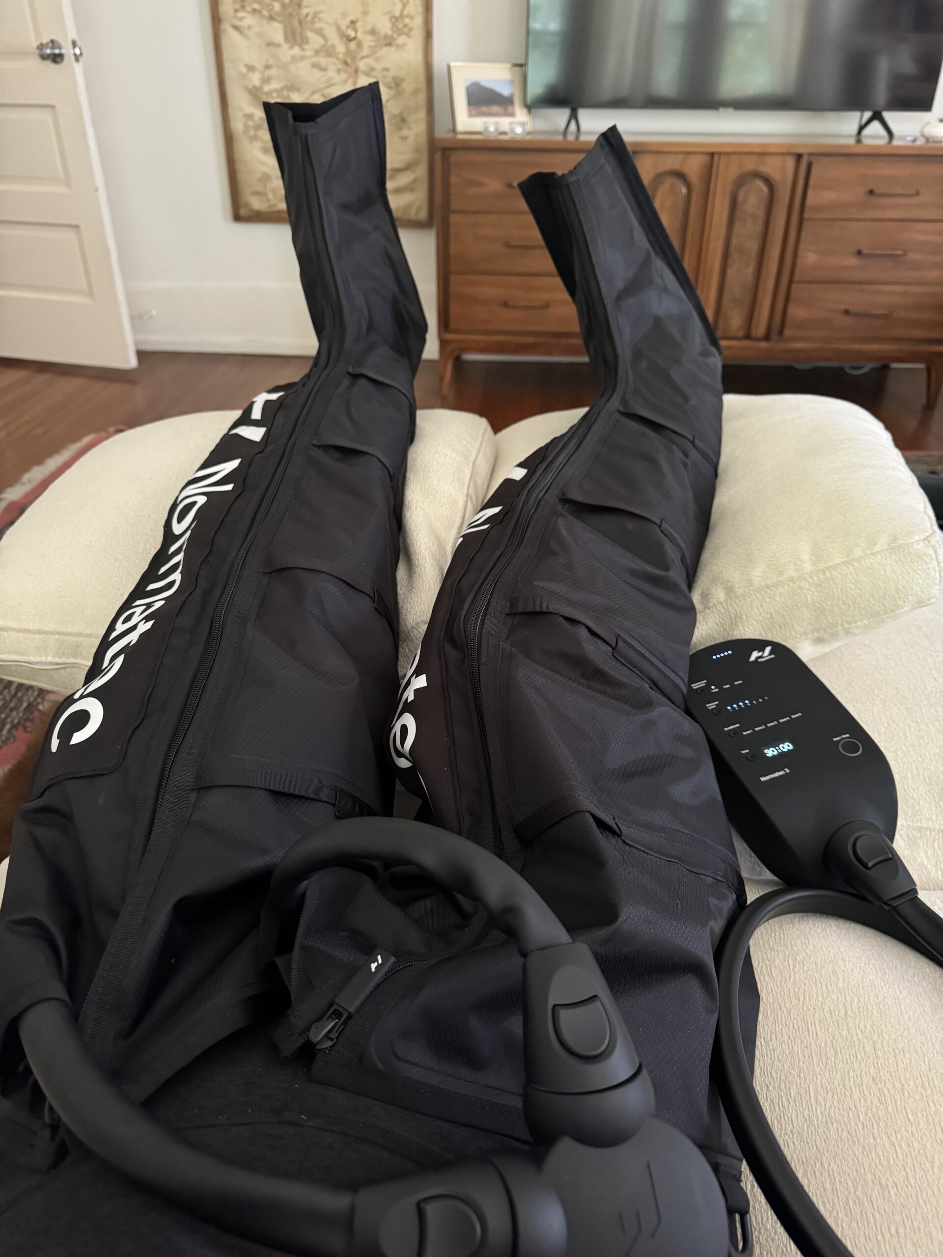 Person using leg compression therapy sleeves while reclining on a couch, with a control device connected to the sleeves and a couple of massage guns nearby.