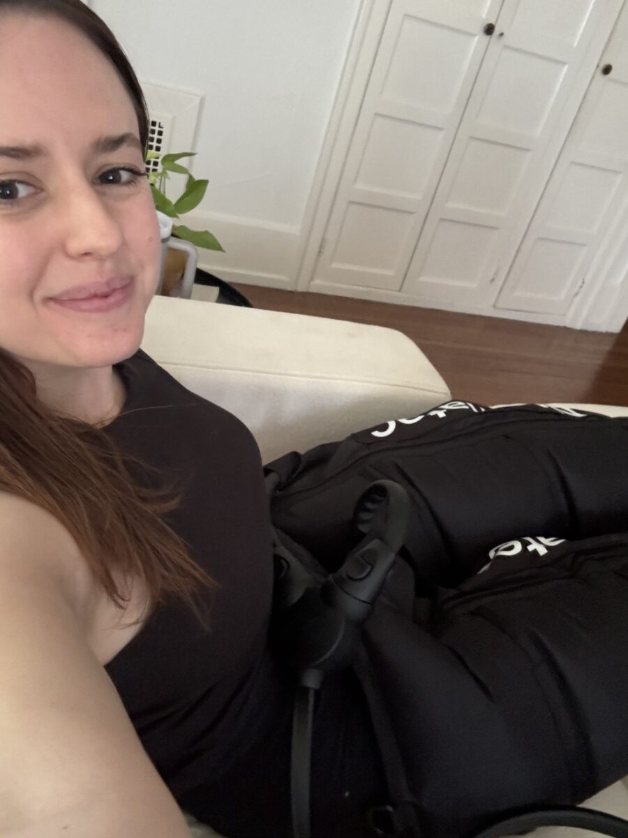 A person wearing a black shirt is laying down on a couch, smiling at the camera, while using black therapy compression sleeves and a massage gun. A plant and a set of white cabinets are visible in the background.