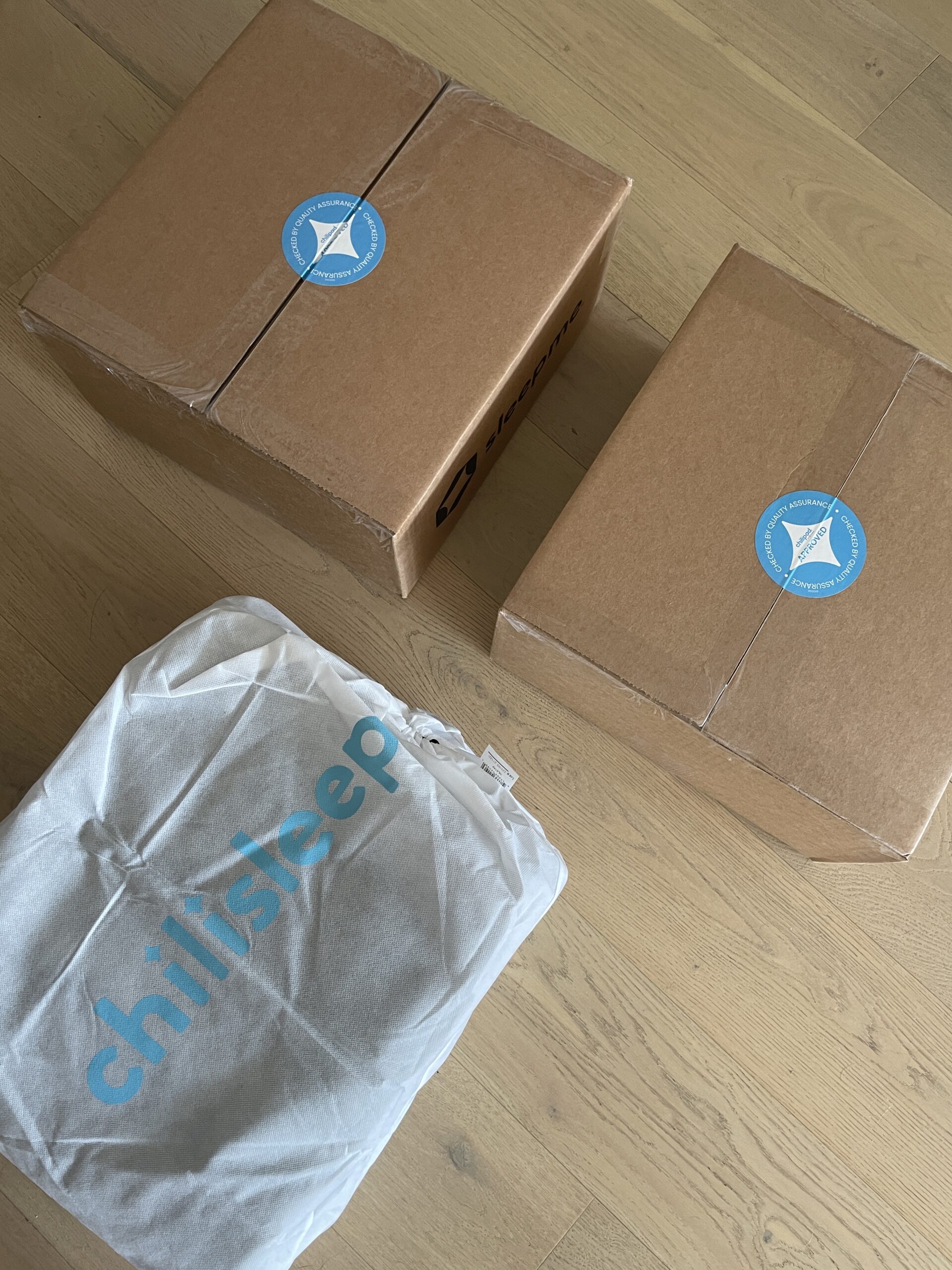 Three packages sit on a wooden floor: two brown cardboard boxes sealed with blue informational stickers and one white fabric package labeled "chilisleep.