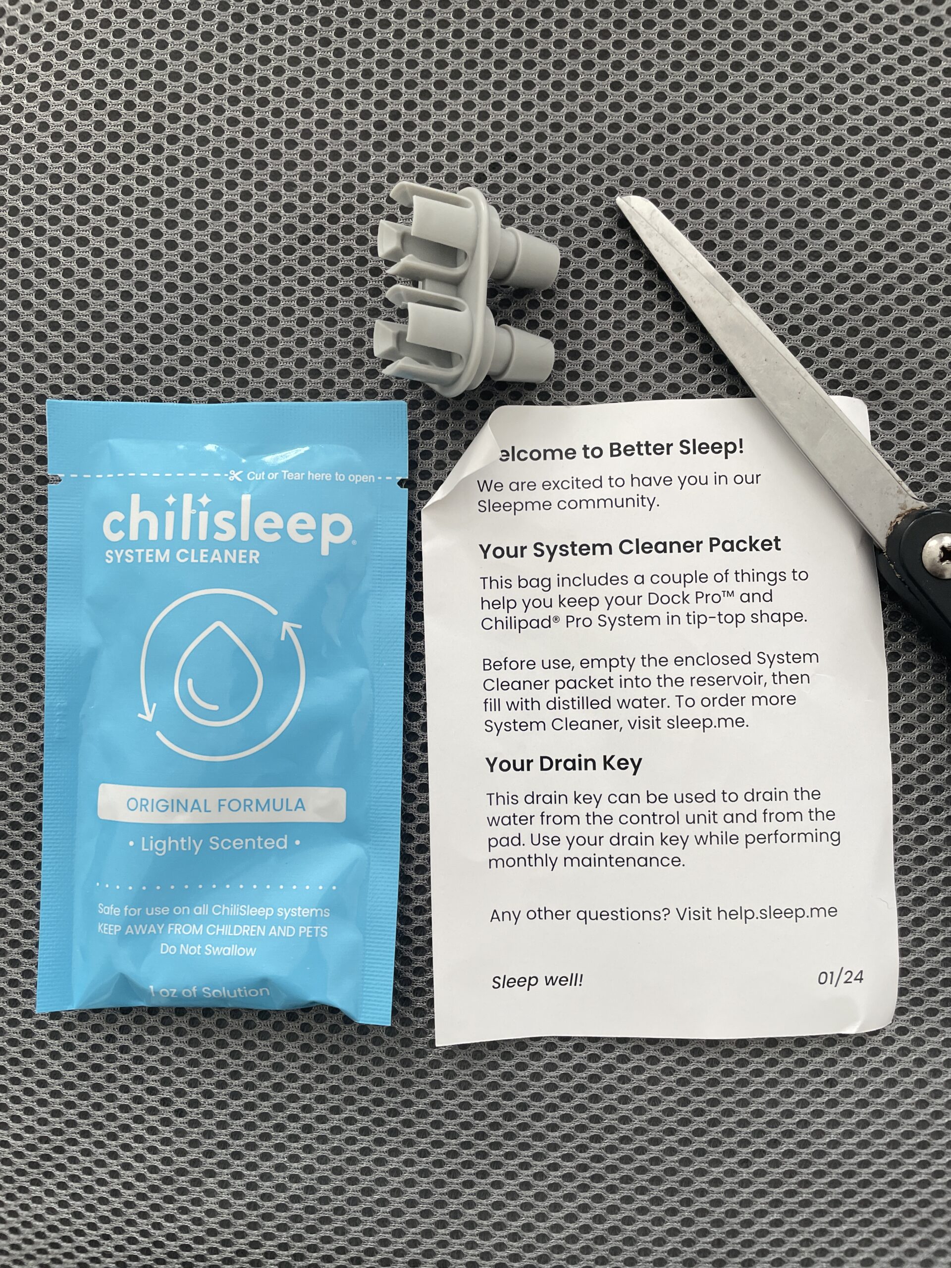 A Chilisleep System Cleaner packet, a paper leaflet with system instructions, and a gray plastic drain key on a gray surface. A pair of scissors is partially visible on the right.