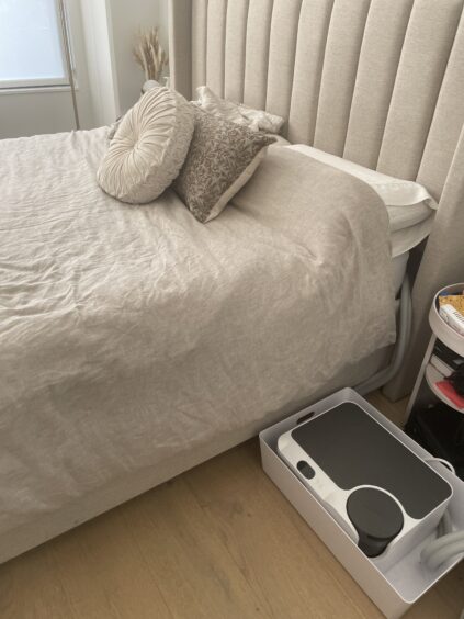 A neatly made bed with beige bedding and decorative pillows. Near the bed on the floor, there is a white tray holding a black and white scale and other items.