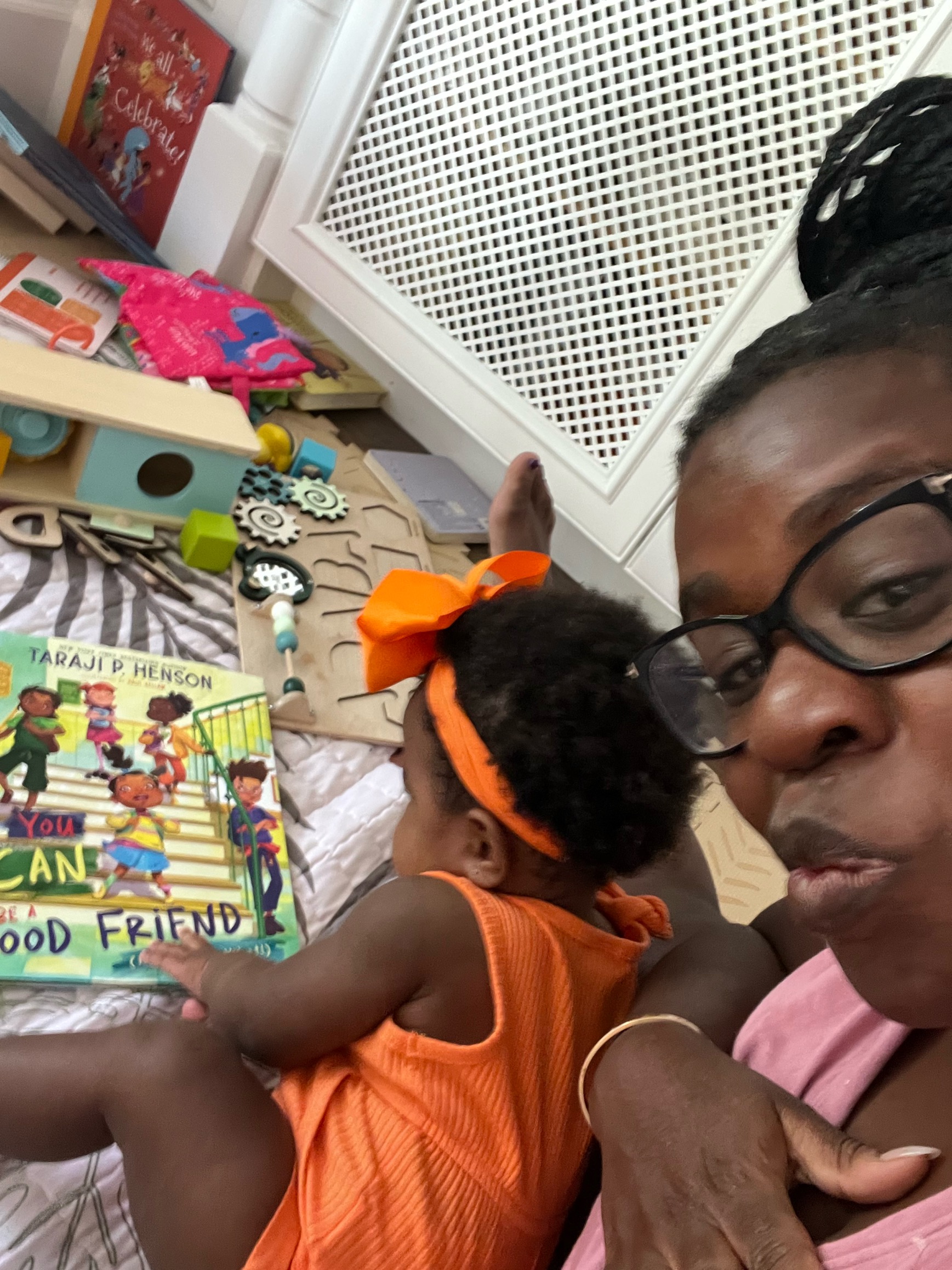 A person wearing glasses holds a child dressed in orange in front of a book and scattered toys on the floor.