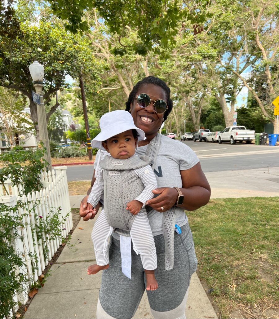 A person smiles while carrying a baby in a front-facing carrier. They are outdoors on a tree-lined street.