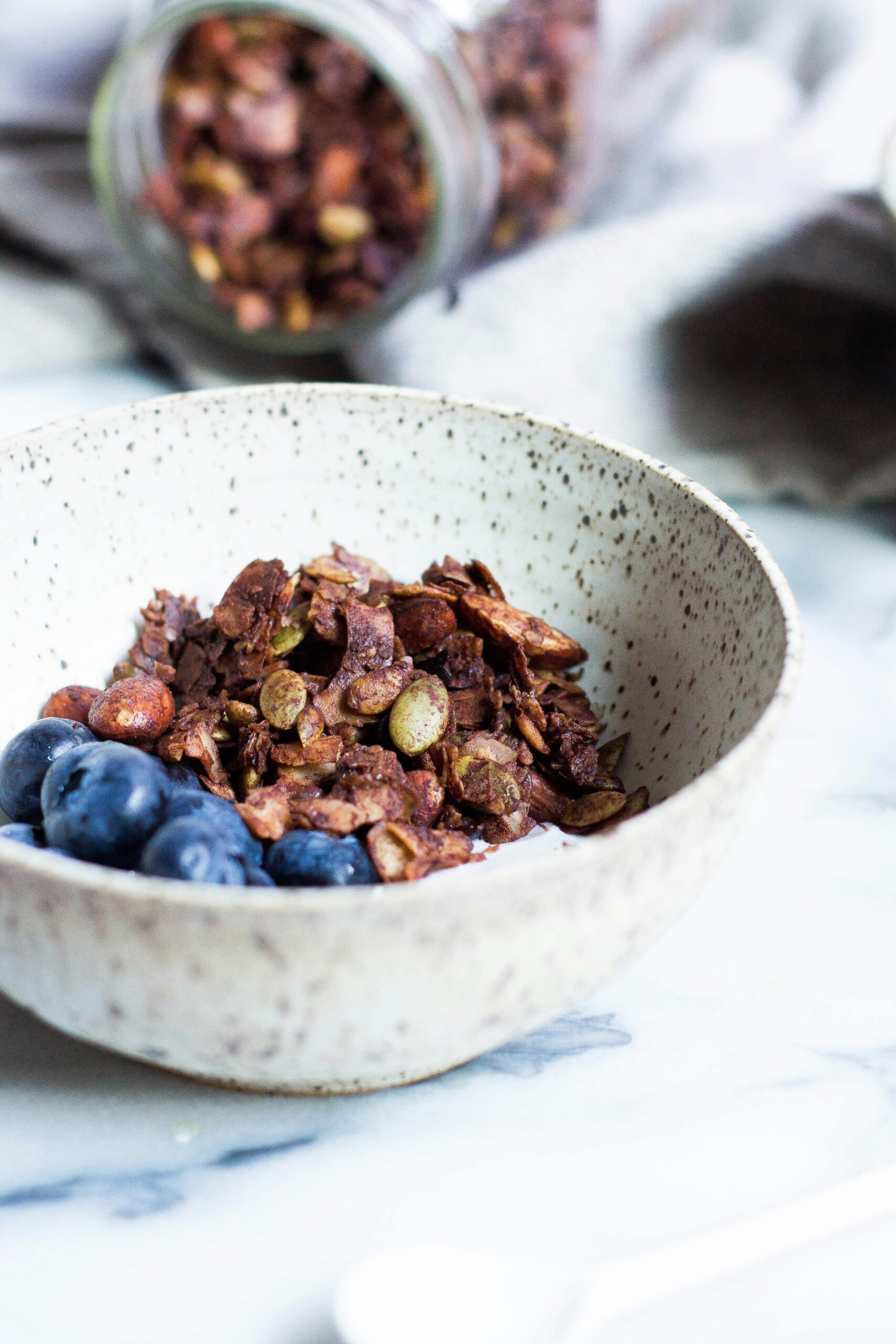 A ceramic bowl of granola and blueberries is on a marble surface, with a jar of granola spilled in the background.