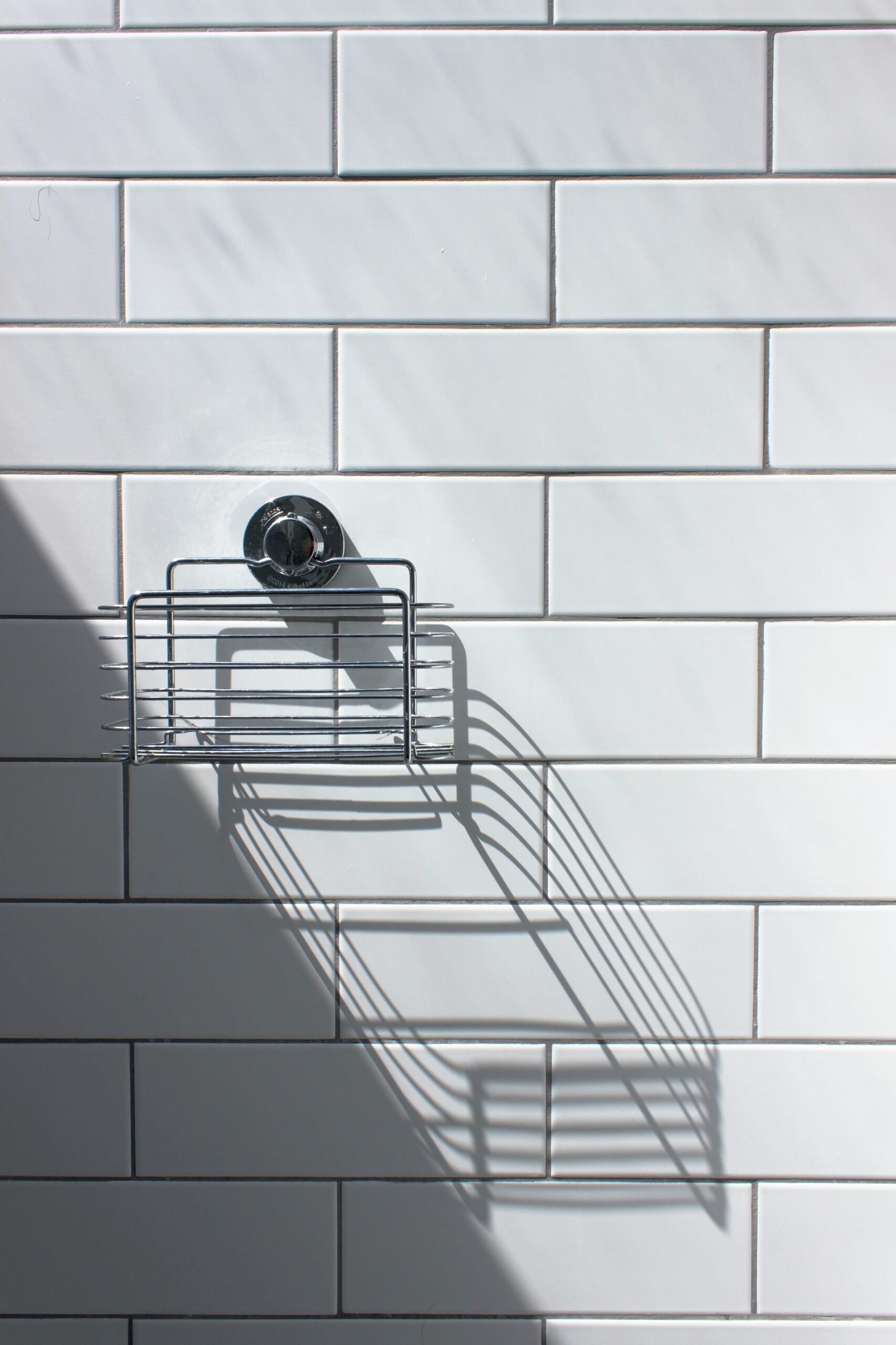 Metal shower caddy mounted on a white tile wall with a long shadow cast from the sunlight.