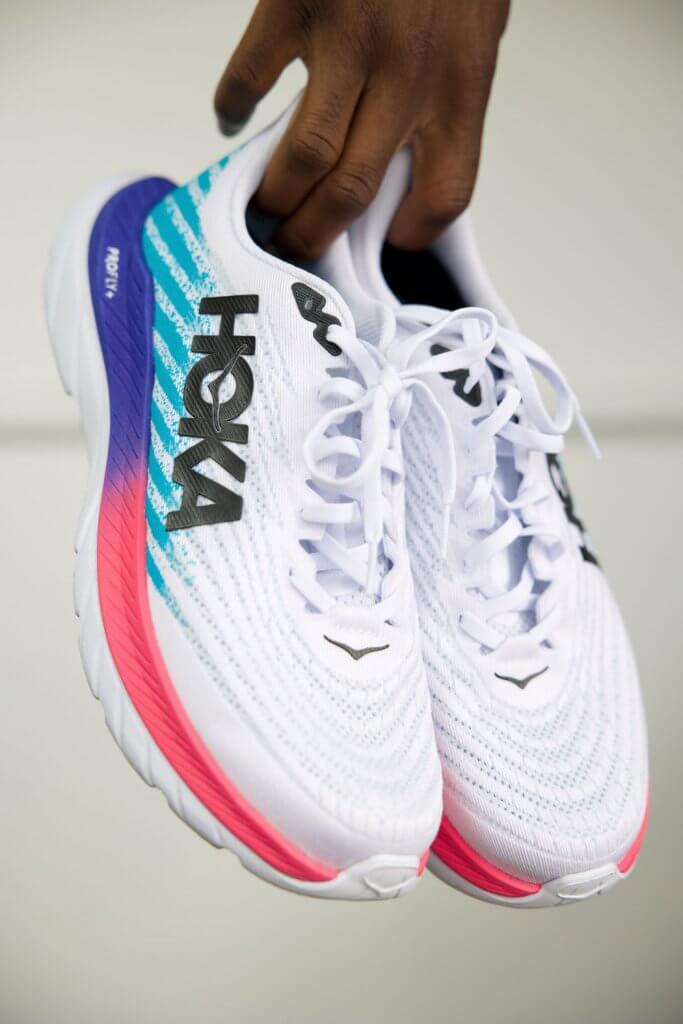 A person holding a pair of white running shoes with blue, pink, and purple accents, and the brand name "HOKA" displayed prominently on the side.