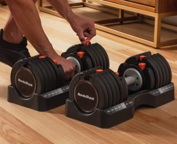 Person adjusting the weights on a pair of NordicTrack adjustable dumbbells placed on a stand on a wooden floor, with massage guns neatly organized in the background.