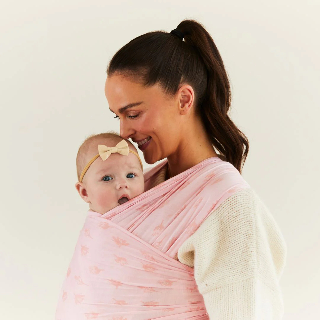 A woman with a ponytail, wearing a white sweater, holds a baby wrapped in a pink cloth. The baby, wearing a headband with a bow, is looking at the camera.